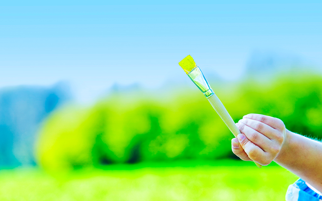 Hand holding a paintbrush with green paint against a natural background of grass and sky