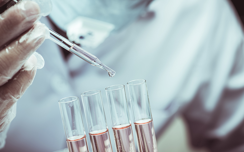 Scientist wearing a lab coat and gloves adding drops using a pipette to four test tubes containing pink liquids.